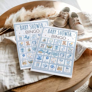 May include: Two "Baby Shower Bingo" cards featuring blue illustrations of baby-related items such as overalls, shoes, and a teddy bear. Beige baby shoes are placed above the cards on a wooden table.