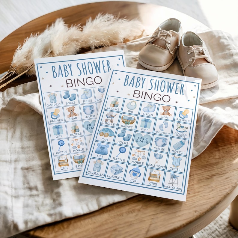 May include: Two "Baby Shower Bingo" cards featuring blue illustrations of baby-related items such as overalls, shoes, and a teddy bear. Beige baby shoes are placed above the cards on a wooden table.