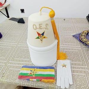 Puede incluir: Un gorro fez de fieltro blanco con una borla amarilla y una estrella bordada en oro con las letras "O.E.S." en pedrería. También se incluye un guante blanco con una estrella bordada en oro y una banda blanca y multicolor con una estrella bordada en oro.