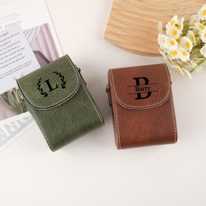 Peut inclure: Deux étuis en cuir, un vert olive et un marron, avec des coutures blanches. L'étui vert porte la lettre noire "L" dans une couronne de laurier. L'étui marron porte le nom "Barry" avec un "D" stylisé au-dessus et en dessous.