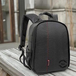 Peut inclure: Sac à dos noir pour appareil photo avec un détail de couture rouge verticale. Le sac à dos a des bretelles rembourrées, une poignée supérieure et une poche latérale en filet. Le nom "Kai" est présent dans le coin inférieur droit.