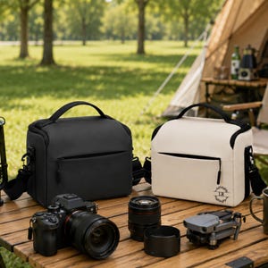 May include: Two rectangular camera bags, one black and one off-white, sit on a wooden table. The off-white bag features a small circular logo with the letters LR inside a leaf wreath. A camera with a lens, an extra lens, and a drone are also present.