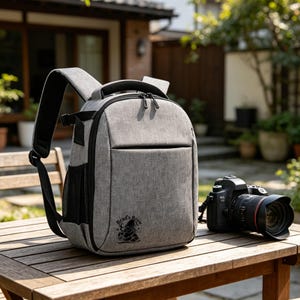 May include: A gray camera backpack with black trim and a top handle, featuring a small black graphic of a gnome holding a mug with the text "Brad's Brew" on the lower front. A black DSLR camera with a lens is positioned next to the backpack.