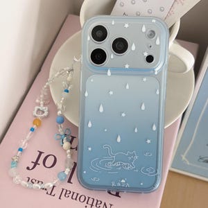 May include: A light blue gradient phone case featuring white raindrop and star icons, a line-art cat in a puddle, and the word "rain" at the bottom. Attached is a beaded phone strap with clear, blue, and white beads, a small white cat charm, and a blue bow.