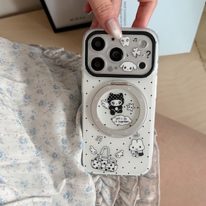 May include: A clear phone case featuring a black polka dot pattern and various black and white cartoon graphics including a winged character, a handbag, a rabbit, and a tooth. A circular magnetic ring stand is centred on the back of the case.
