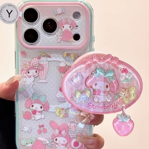 Puede incluir: Una funda de teléfono rosa y transparente con un soporte a juego. La funda presenta personajes de dibujos animados, fresas y dulces. El soporte del teléfono tiene forma ovalada y tiene la palabra "Strawberry".