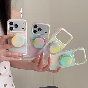 May include: Four clear smartphone cases with pastel gradient circular grips. Each case features a rectangular camera cutout and a soft, blended color transition of yellow, pink, blue, and green hues across the back and the circular grip attachment.