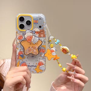 Puede incluir: Funda de teléfono transparente con un marco de cámara amarillo y un diseño de dibujos animados con personajes durmientes, estrellas y lunas. La funda tiene un pop socket en forma de estrella y una correa de cuentas con dijes. El texto "Twinkle Twinkle" es visible.