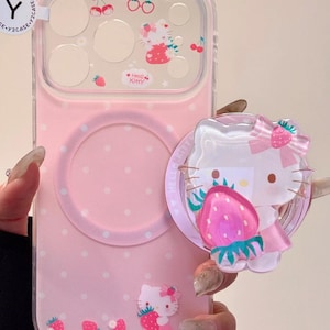 May include: Pink phone case with a Hello Kitty design. The case features a clear camera lens protector with cherry and Hello Kitty graphics. A matching pop socket with a strawberry and Hello Kitty is attached. The case has a polka dot pattern and strawberry accents.