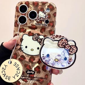 Puede incluir: Una funda transparente para teléfono con diseño de estampado de leopardo y temática de Hello Kitty. La funda tiene un charm a juego con un diseño de Hello Kitty y la palabra "Lucky". La funda está diseñada para un teléfono.