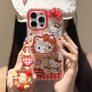 May include: Clear phone case with a red border and a red and white gingham pattern. The case features Hello Kitty graphics, including a chef and a bow. A beaded wrist strap with red, yellow, and white beads is attached.