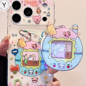 May include: Clear phone case with colorful cartoon graphics and the text "Tomago Tchi". The case features various cute characters, stars, and musical notes. A matching charm is attached.
