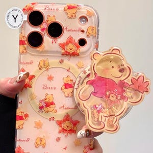 Puede incluir: Funda transparente para teléfono con diseño de Winnie the Pooh, hojas de otoño y la palabra "Autumn". La funda tiene un popsocket a juego con un gráfico de Winnie the Pooh. La funda está sujeta por una persona.