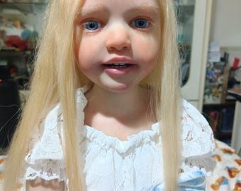 Puede incluir: Una muñeca realista con piel clara, ojos azules y cabello rubio largo. La muñeca lleva un vestido de encaje blanco con detalles florales azules. La cara de la muñeca está finamente detallada, creando una apariencia realista.