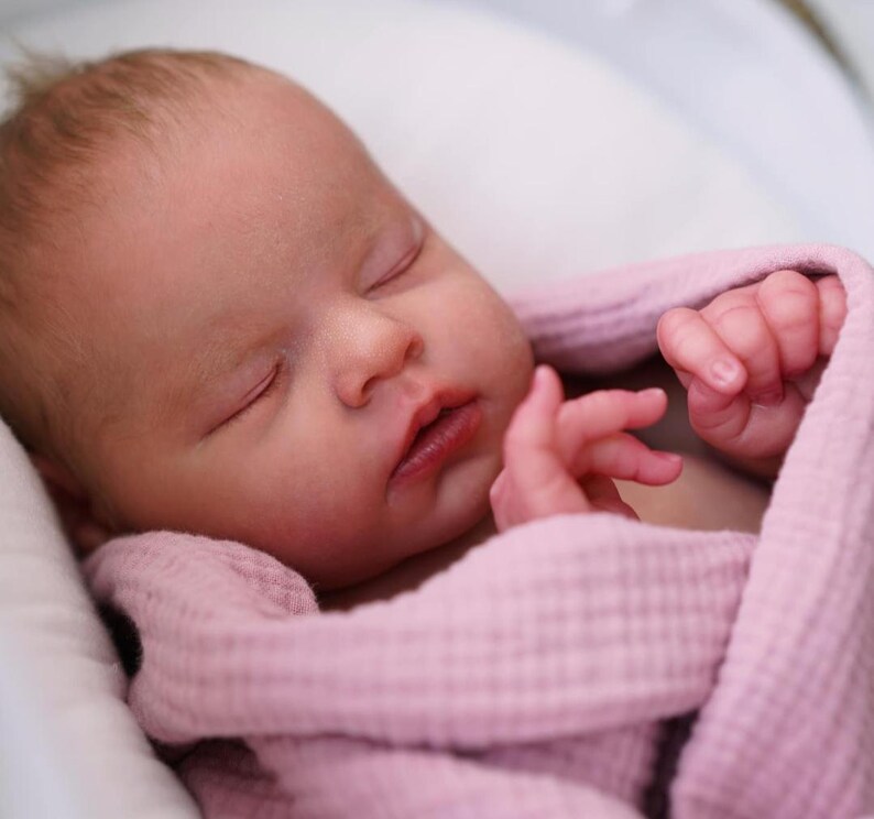 May include: A newborn baby sleeping peacefully, wrapped in a soft, light pink blanket. The baby's delicate features are visible, with closed eyes and small hands. The baby is nestled in a white, cushioned surface.