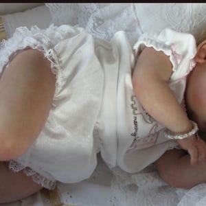 May include: A lifelike baby doll with fair skin and closed eyes, dressed in a white onesie with lace trim. The doll is lying on a white, textured surface, wearing a bracelet. The doll's hair is light brown.