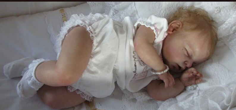 May include: A lifelike baby doll with fair skin and closed eyes, dressed in a white onesie with lace trim. The doll is lying on a white, textured surface, wearing a bracelet. The doll's hair is light brown.
