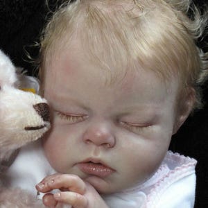 May include: A lifelike baby doll with closed eyes and light blonde hair. The doll is wearing a white outfit with pink trim and is holding a stuffed animal. The doll's skin has a realistic tone.