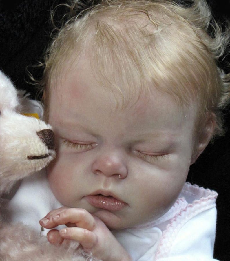 May include: A lifelike baby doll with closed eyes and light blonde hair. The doll is wearing a white outfit with pink trim and is holding a stuffed animal. The doll's skin has a realistic tone.