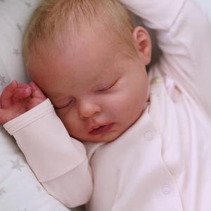 May include: A sleeping baby with fair skin and light blonde hair, wearing a light pink onesie. The baby is lying on a white pillow with a star pattern. The baby's arms are raised above its head.