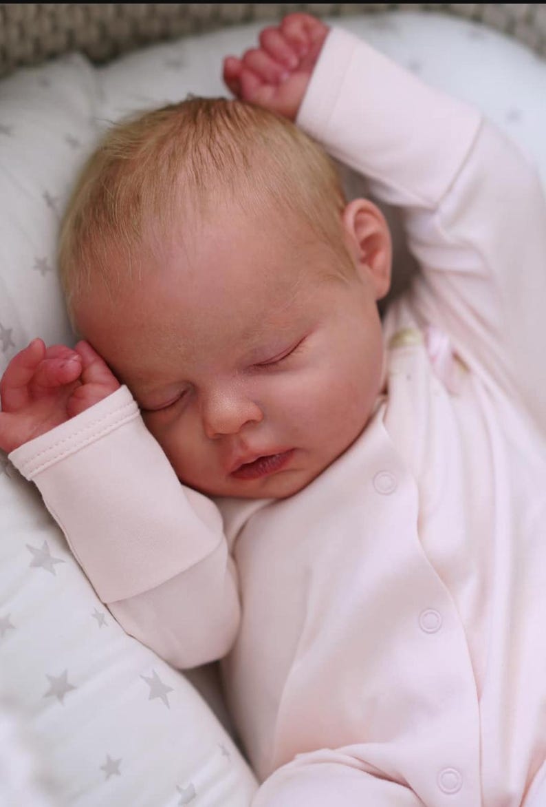 May include: A sleeping baby with fair skin and light blonde hair, wearing a light pink onesie. The baby is lying on a white pillow with a star pattern. The baby's arms are raised above its head.