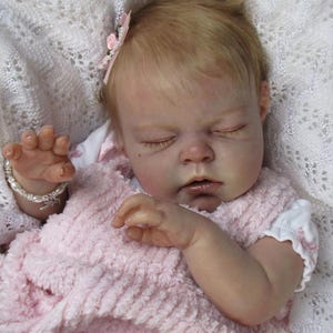 May include: A lifelike baby doll with closed eyes, wearing a pink textured dress and a white undershirt. The doll has blonde hair and a pink flower hair accessory. A pearl bracelet adorns the doll's wrist.