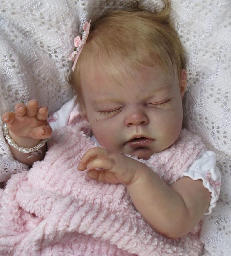 May include: A lifelike baby doll with closed eyes, wearing a pink textured dress and a white undershirt. The doll has blonde hair and a pink flower hair accessory. A pearl bracelet adorns the doll's wrist.