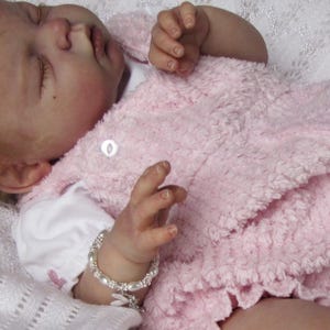 May include: A lifelike baby doll wearing a pink textured dress and a white shirt. The doll has a pink bow in its hair and a silver bracelet. The doll is lying on a white, textured blanket. The doll is wearing white shoes with pink socks.