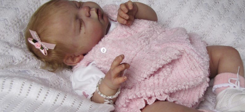 May include: A lifelike baby doll wearing a pink textured dress and a white shirt. The doll has a pink bow in its hair and a silver bracelet. The doll is lying on a white, textured blanket. The doll is wearing white shoes with pink socks.