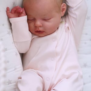 May include: A sleeping baby wearing a light pink onesie with button closures. The baby is lying on a star-patterned pillow. The baby's arms are raised above its head.