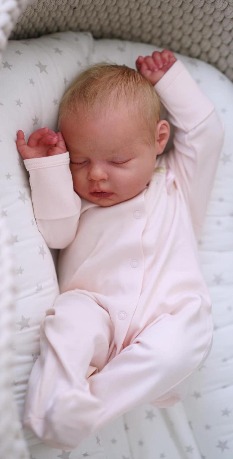 May include: A sleeping baby wearing a light pink onesie with button closures. The baby is lying on a star-patterned pillow. The baby's arms are raised above its head.
