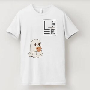 以下が含まれることがあります： カボチャを持った漫画の幽霊が描かれた白いTシャツ。シャツには「LDK」の文字とロゴが入った黒い正方形もあります。幽霊はベージュ色で黒い目をし、オレンジ色のカボチャを持っています。