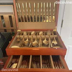 May include: A wooden cutlery chest with a variety of gold-colored silverware. The top section holds knives and serving utensils, while the drawer below contains forks, spoons, and more. The silverware has a shiny, reflective finish.