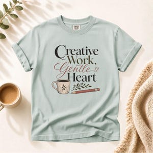 Op de afbeelding: Een licht saliegroen T-shirt met korte mouwen en de zwarte en bordeauxrode tekst Creative Work, Gentle Heart. De afbeelding bevat een beige koffiemok met een bladontwerp, een klein takje met bladeren en een potlood met een roze gum.
