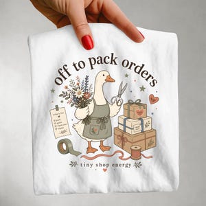 Op de afbeelding: Een wit t-shirt met een afbeelding van een gans in een schort, die een boeket bloemen en een schaar vasthoudt. Het ontwerp bevat een stapel van drie cadeauverpakkingen, een rol tape, een lijst met de tekst today's list, en de tekst off to pack orders en tiny shop energy.