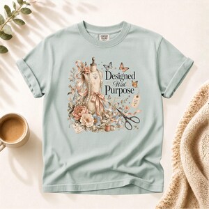 Op de afbeelding: Een licht saliegroen t-shirt met een grafische print van een paspop, bloemstukken, vlinders, een schaar en een klosje garen. De tekst "Designed With Purpose" is in het zwart in het midden van het ontwerp gedrukt.