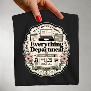 Op de afbeelding: Een zwart t-shirt met een afbeelding met de tekst "Everything Department", "Quietly keeping it together" en "With excellent taste". Het ontwerp bevat een laptop, kalender, map, potlood en iconen voor admin-professionals, online reviewers, AI-testers en kleine ondernemers, omringd door bloemmotieven.