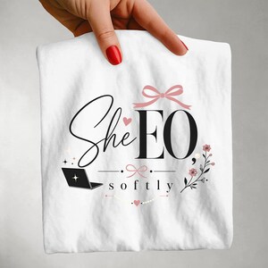 Op de afbeelding: Een wit t-shirt met een zwart en roze grafisch ontwerp. De tekst luidt SheEO softly. Het ontwerp bevat een zwart laptopicoon, een roze strik, kleine roze hartjes en een bloemtakje. De belettering combineert script- en schreeflettertypes.