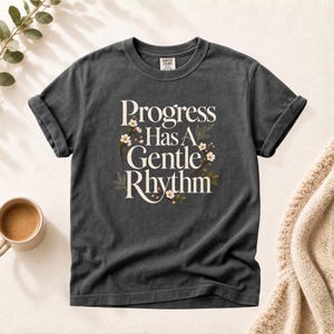 Op de afbeelding: Een antracietgrijs t-shirt met korte mouwen en de witte tekst "Progress Has A Gentle Rhythm" in het midden op de borst. De tekst is omringd door delicate illustraties van kleine witte bloemen en dunne bruine takjes met bladeren.