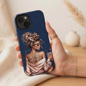 Op de afbeelding: Marineblau telefoonhoesje met een illustratie van een vrouw met een luipaardprint strik, die een koffiekopje vasthoudt. De hoes heeft de tekst "Be Real not perfect" en wordt vastgehouden in een hand met gouden ringen en een armband.