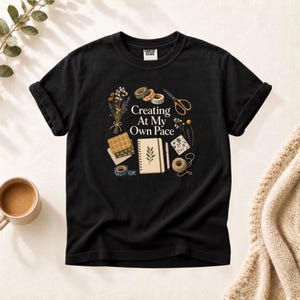 Op de afbeelding: Een zwart t-shirt met korte mouwen en een grafische print met de tekst "Creating At My Own Pace", omringd door illustraties van een notitieboek, een schaar, washi-tape, droogbloemen, stofstalen, een pen en een bolletje touw.