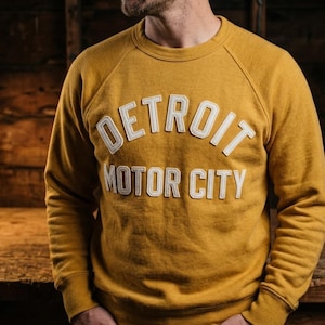 以下が含まれることがあります： 白のブロック文字で「DETROIT MOTOR CITY」と書かれたマスタードイエローのスウェットシャツ。スウェットシャツはクルーネックとラグランスリーブです。木製の背景の前に人が立っています。