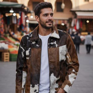 May include: A brown, black, and white cowhide jacket with a zipper closure. The jacket has a collar and is worn over a white t-shirt. The person is wearing dark jeans. The background is a marketplace.