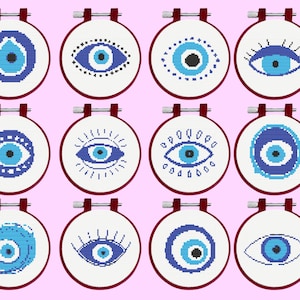 Puede incluir: Doce bastidores de bordado circulares con tela blanca que presentan diseños de ojos en punto de cruz en varios tonos de azul, negro y blanco. Cada bastidor tiene un tornillo de metal en la parte superior y un borde rojo oscuro.