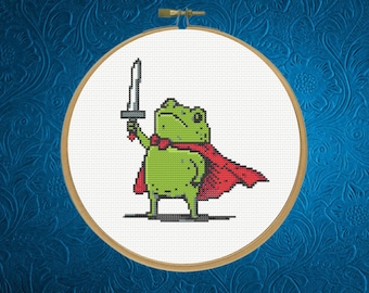 May include: A cross-stitch embroidery piece in a circular wooden hoop featuring a pixelated green frog wearing a red cape and a red bowtie, holding a grey sword aloft. The design is stitched on a white fabric background with a visible grid pattern.