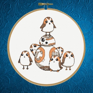 May include: Cross-stitch embroidery of Star Wars characters BB-8 and Porgs. The design features BB-8 in the center, surrounded by several Porgs. The embroidery is set in a round wooden hoop against a white background, with a blue floral patterned background.