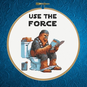 May include: A white cross-stitch embroidery hoop with a brown Wookiee character sitting on a toilet and reading a book. The text "USE THE FORCE" is above the character.