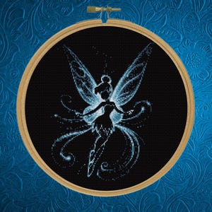 May include: A circular wooden embroidery hoop containing a black fabric piece with a cross-stitched design of a fairy. The fairy is rendered in shades of light blue and white, featuring large wings and swirling trails of light against the dark background.