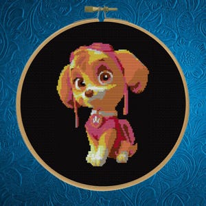 May include: A circular wooden embroidery hoop containing a cross-stitch pattern of a cartoon puppy with tan and yellow fur, wearing a pink aviator hat and a pink vest with a silver badge, set against a solid black background.