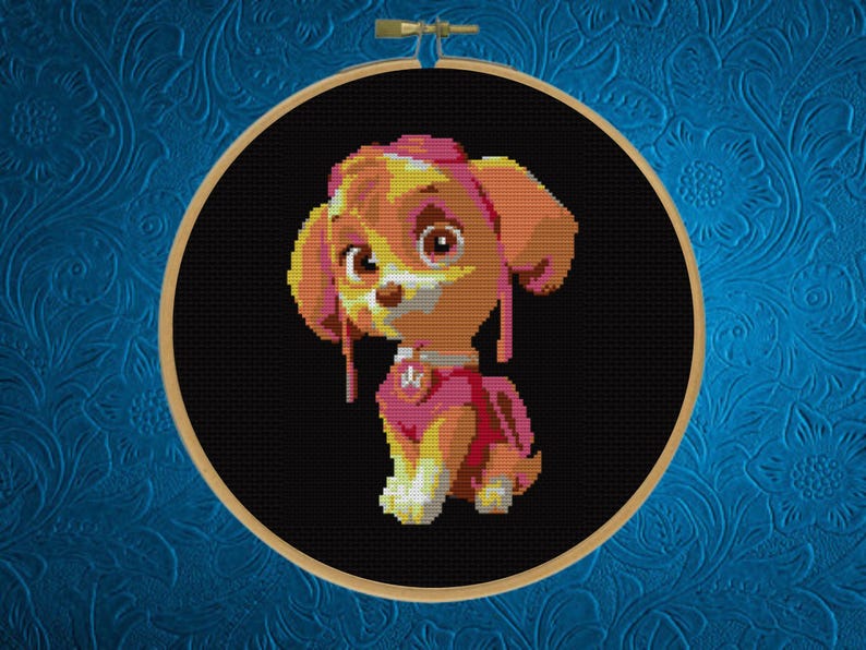 May include: A circular wooden embroidery hoop containing a cross-stitch pattern of a cartoon puppy with tan and yellow fur, wearing a pink aviator hat and a pink vest with a silver badge, set against a solid black background.